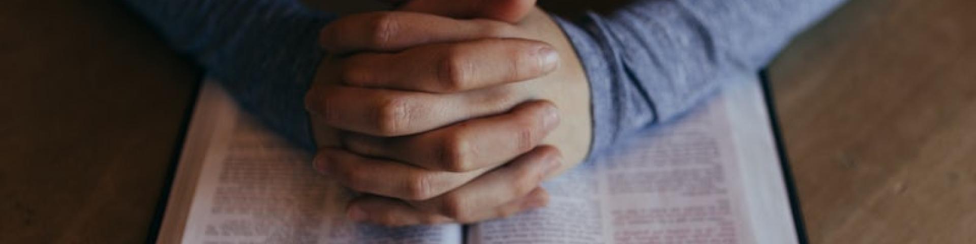 A person's hands folded in prayer on top of a bible
