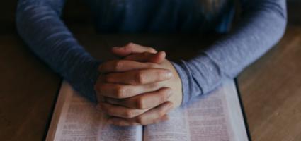 A person's hands folded in prayer on top of a bible
