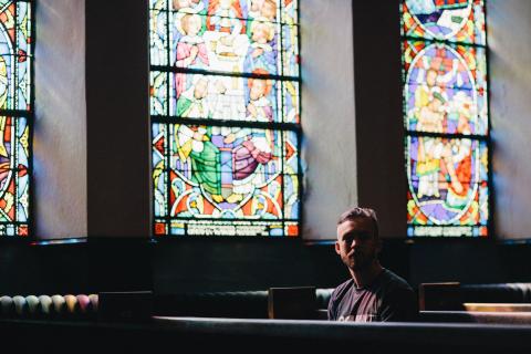 Picture of young adult male sitting in a pew in a dark church with stained glass weekends in the background 